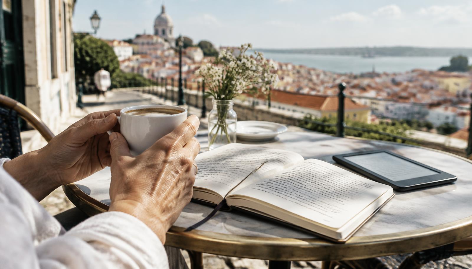 Hands holding coffee beside an open notebook and an e-reader at a café table